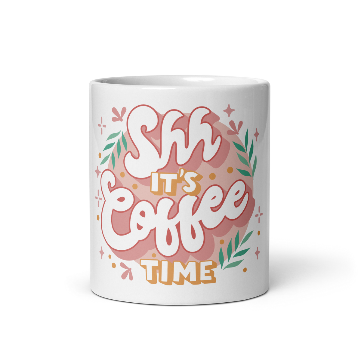 A white ceramic mug featuring a colorful graphic design with the text 'Shh It's Coffee Time' in stylized pink, white, and orange lettering, surrounded by green leaves and small decorative stars.