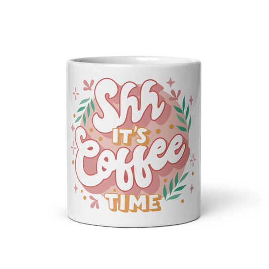 A white ceramic mug featuring a colorful graphic design with the text 'Shh It's Coffee Time' in stylized pink, white, and orange lettering, surrounded by green leaves and small decorative stars.